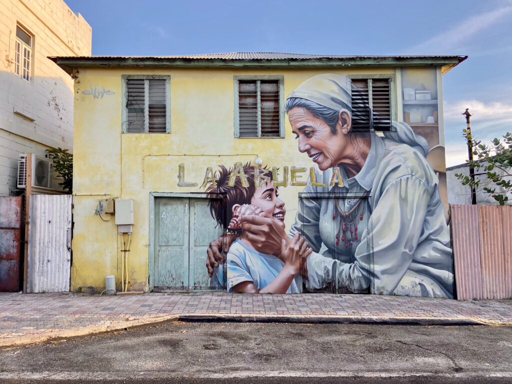 Mural La Abuela by Chemis, painted in San Nicolas, Aruba, 2025: an elderly woman and a child on a yellow house wall.