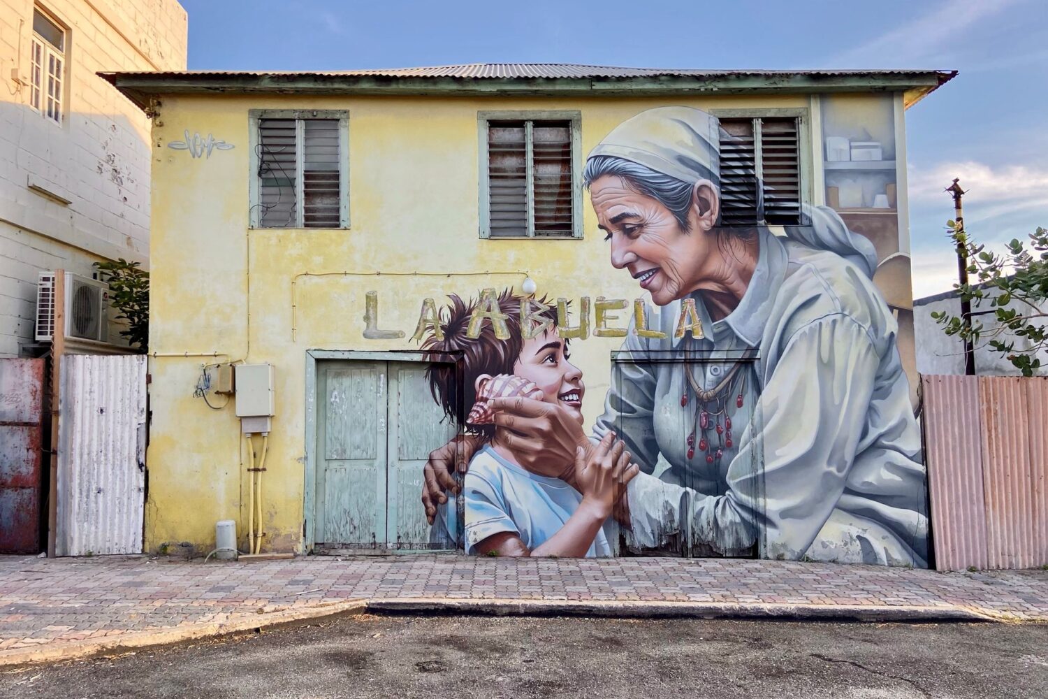 Mural La Abuela by Chemis, painted in San Nicolas, Aruba, 2025: an elderly woman and a child on a yellow house wall.