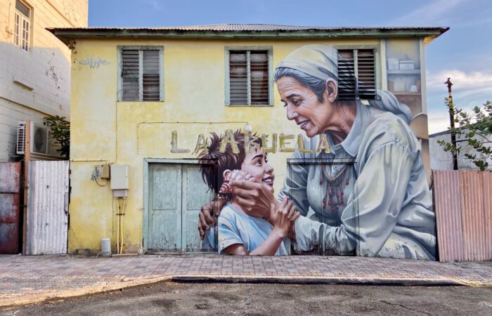 Mural La Abuela by Chemis, painted in San Nicolas, Aruba, 2025: an elderly woman and a child on a yellow house wall.