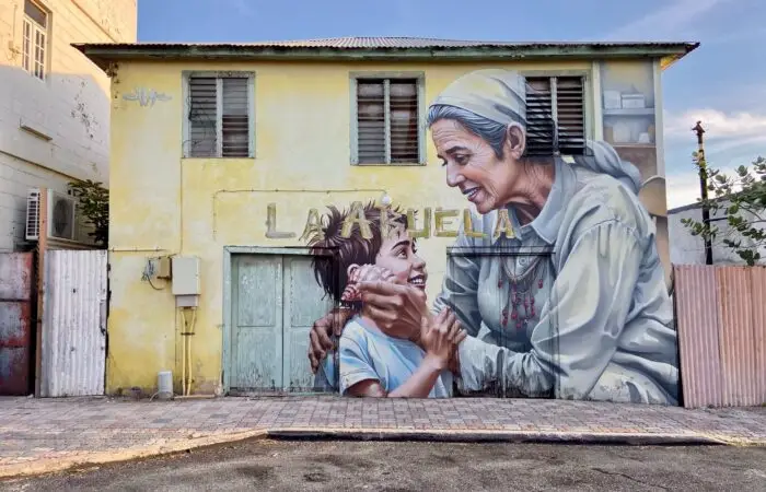 Mural La Abuela by Chemis, painted in San Nicolas, Aruba, 2025: an elderly woman and a child on a yellow house wall.
