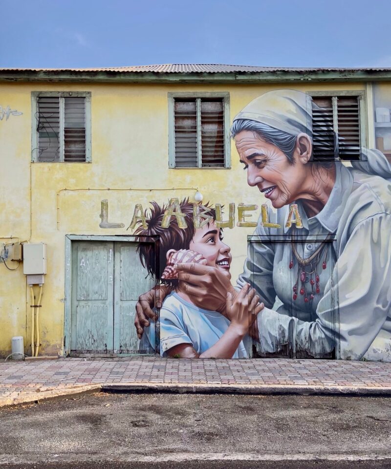 Mural La Abuela by Chemis, painted in San Nicolas, Aruba, 2025: an elderly woman and a child on a yellow house wall.