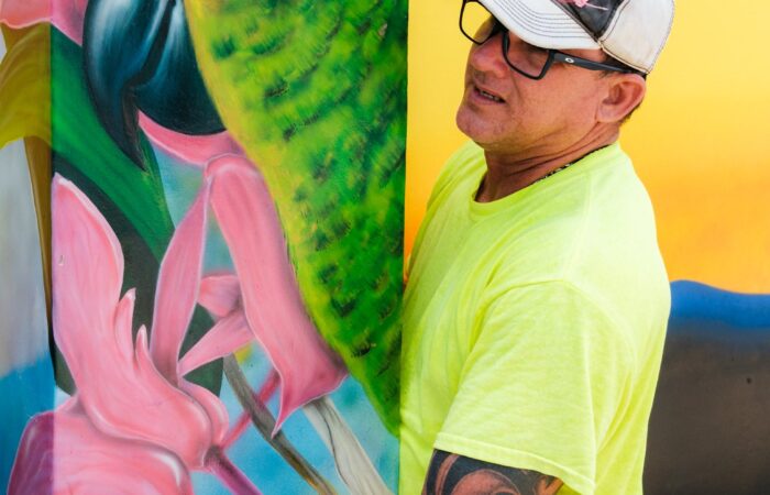 An artist standing beside a finished parrot mural during the Aruba Mural Festival 2025 in San Nicolas.