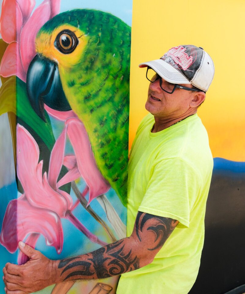 An artist standing beside a finished parrot mural during the Aruba Mural Festival 2025 in San Nicolas.