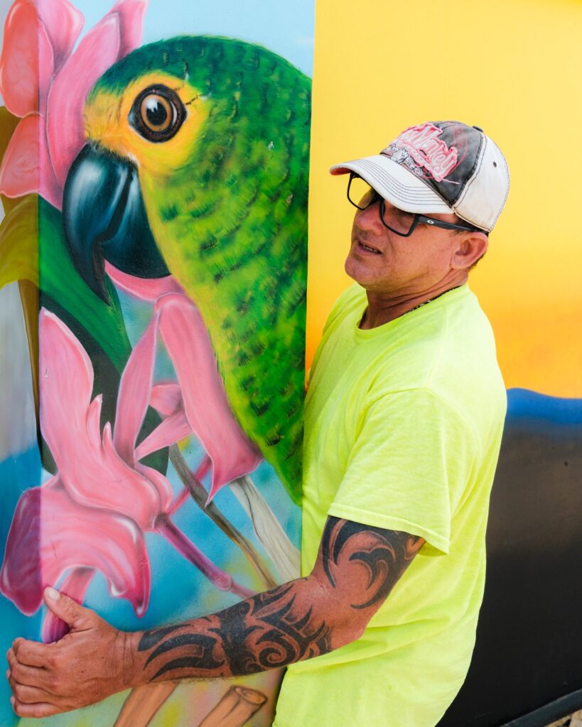 An artist standing beside a finished parrot mural during the Aruba Mural Festival 2025 in San Nicolas.