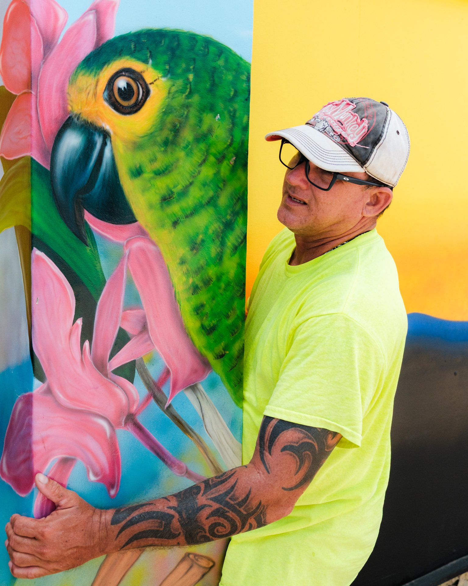 An artist standing beside a finished parrot mural during the Aruba Mural Festival 2025 in San Nicolas.